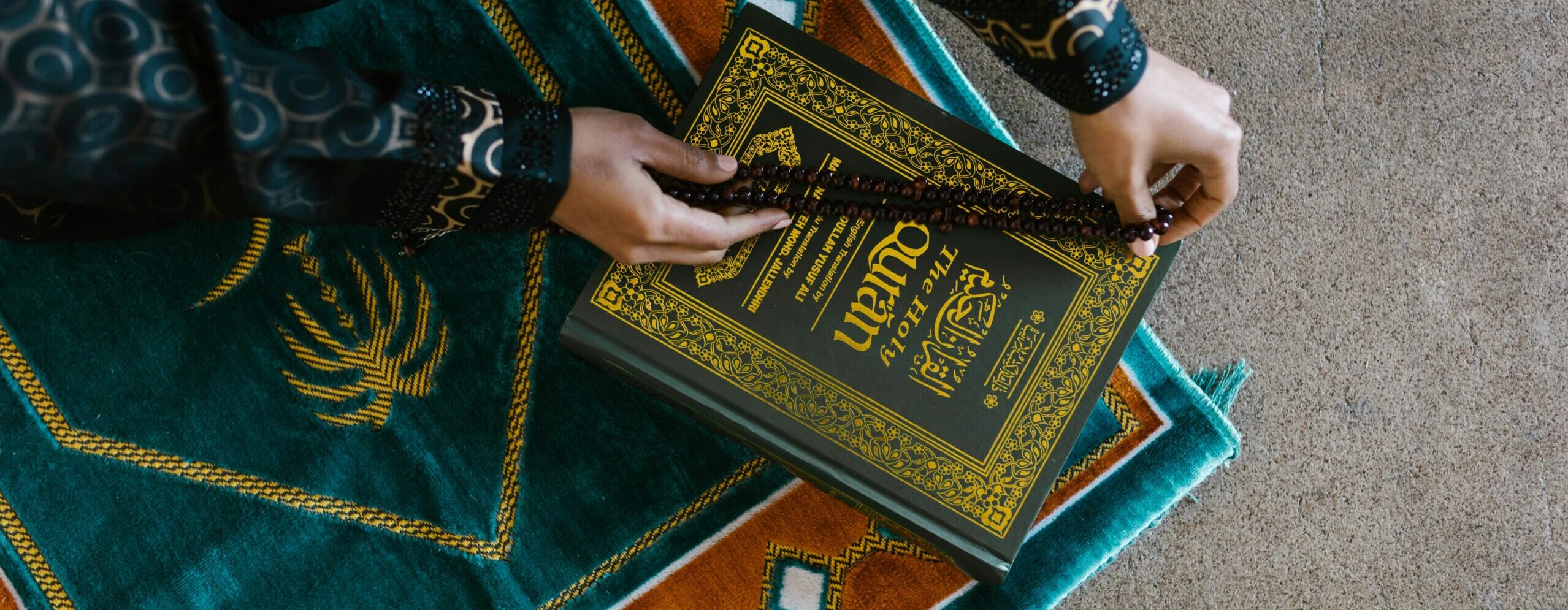 Top view of a person holding prayer beads and Quran on a colorful rug, symbolizing Islamic faith and worship.